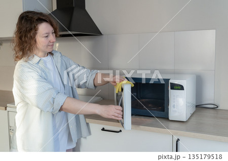 Woman wipes a white microwave with a yellow cloth. Performing everyday household chores and maintaining the cleanliness of kitchen appliances Woman wipes a white microwave with a yellow cloth. Performing everyday household chores and maintaining the cleanliness of kitchen appliances 135179518