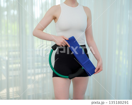 Young woman holding fitness ring and resistance band at home. Concept of home workout, strength training, and healthy lifestyle. Female preparing equipment for online fitness session. Young woman holding fitness ring and resistance band at home. Concept of home workout, strength training, and healthy lifestyle. Female preparing equipment for online fitness session. 135180291