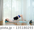 Young woman exercising at home with resistance band on yoga mat. Concept of fitness, healthy lifestyle, and online workout. Female performing glute bridge while following virtual training on tablet. 135180293