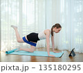 Young woman exercising at home on yoga mat with resistance band. Concept of fitness, strength training, and online workout. Female performing leg raise while following virtual fitness class on tablet. 135180295