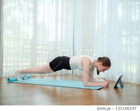 Young woman doing plank exercise at home on yoga mat. Concept of core strength, fitness, and online workout. Female holding plank position while following virtual fitness class on a tablet. Young woman doing plank exercise at home on yoga mat. Concept of core strength, fitness, and online workout. Female holding plank position while following virtual fitness class on a tablet. 135180297