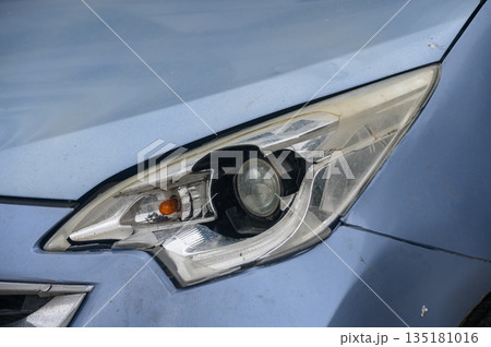 Detailed close-up of a blue car's headlight showcasing intricate design and wear near a busy urban street in bright daylight 135181016