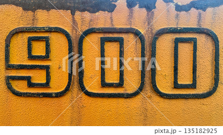Close-up of industrial number 900 on a weathered yellow textured metal surface. Close-up of industrial number 900 on a weathered yellow textured metal surface. 135182925