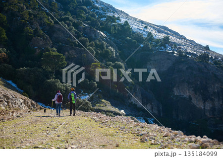 Hikers On a Mountain Trail With Snow Patches, Forest, and Rocky Cliffs Exploring Outdoors 135184099