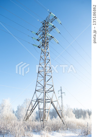 A tall power transmission tower rises in snow-covered landscape under clear blue sky. Frost covers metal parts of the structure A tall power transmission tower rises in snow-covered landscape under clear blue sky. Frost covers metal parts of the structure 135186862