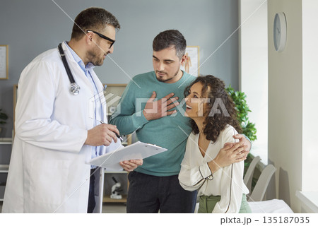 Smiling happy young couple talking with a male family doctor therapist in medical clinic. Smiling happy young couple talking with a male family doctor therapist in medical clinic. 135187035