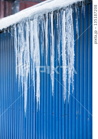 Roof of building with blue color metal wall covered with sharp icicles. Thaw, winter weather 135187208