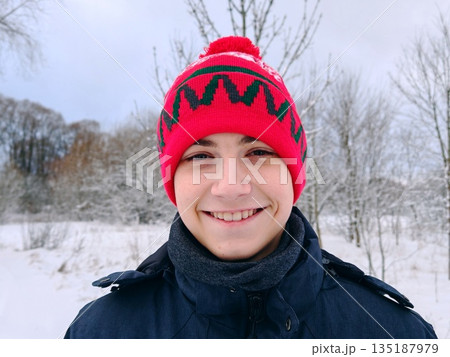 Smiling teenage boy in red winter hat outdoors. Happy young guy in festive red knit beanie with pom-pom and warm jacket against snowy blurred background. Smiling teenage boy in red winter hat outdoors. Happy young guy in festive red knit beanie with pom-pom and warm jacket against snowy blurred background. 135187979