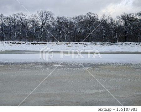 Frozen lake under stormy winter sky. Vast icy lake surface with snow patches, dense bare forest horizon and dramatic dark clouds overhead. 135187983