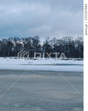 Frozen lake under stormy winter sky. Vast icy lake surface with snow patches, dense bare forest horizon and dramatic dark clouds overhead. 135187984