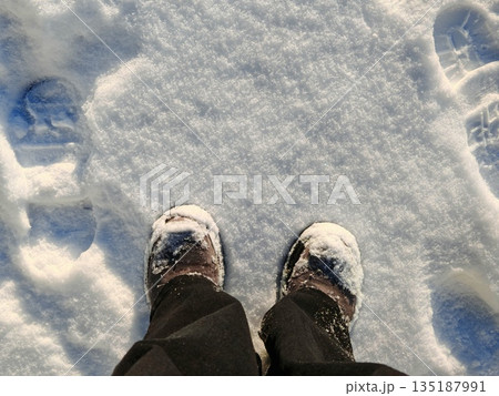 Winter boots standing in fresh snow. POV shot of snowy winter boots with footprints around on bright white snow surface under sunlight. 135187991