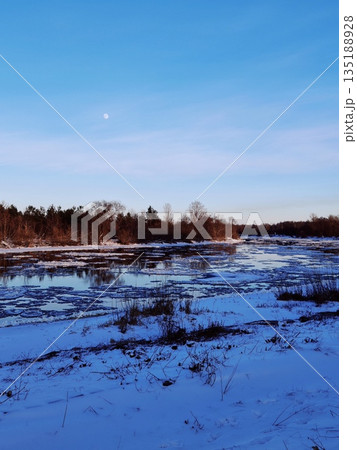 Frozen river reflects bare trees and moon under clear blue winter sky, serene nature landscape. Ice-covered river with snowy banks, forest silhouettes, pale moon in twilight sky, peaceful winter scene Frozen river reflects bare trees and moon under clear blue winter sky, serene nature landscape. Ice-covered river with snowy banks, forest silhouettes, pale moon in twilight sky, peaceful winter scene 135188928