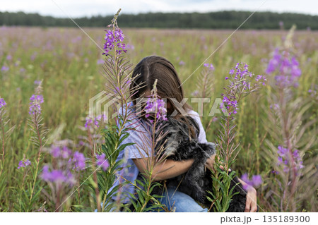 young woman holding gray fluffy senior dog in hands on meadow with flowers of fireweed or great willowherb, pet love 135189300