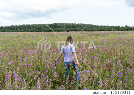 Young beautiful blond woman in purple shirt from behind walking in the meadow among flowers of fireweed Young beautiful blond woman in purple shirt from behind walking in the meadow among flowers of fireweed 135189311