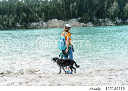 Young woman traveler in cap with backpack walking on sandy shore of blue lake or river with fluffy mixed breed gray dog, active lifestyle, travel with pets Young woman traveler in cap with backpack walking on sandy shore of blue lake or river with fluffy mixed breed gray dog, active lifestyle, travel with pets 135189316