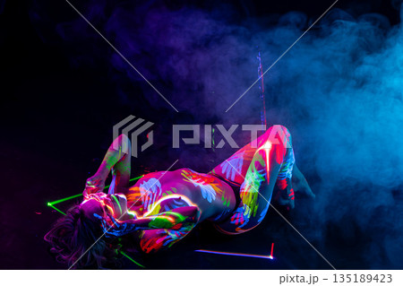 Woman in vibrant UV body paint lies amidst lasers and smoke in a dark studio. Woman in vibrant UV body paint lies amidst lasers and smoke in a dark studio. 135189423