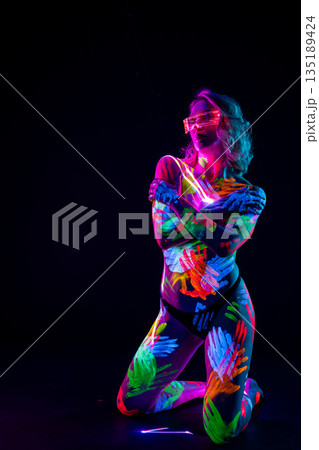 Woman with glowing neon handprints and futuristic glasses kneeling in blacklight on a dark background. 135189424