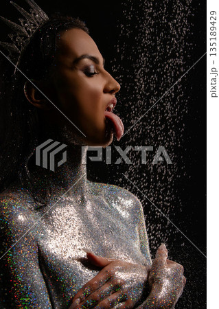 Profile of a sensual woman in a crown and silver glitter body art catching falling water droplets on her tongue. 135189429
