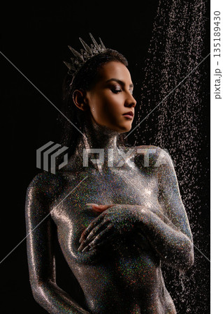 Regal woman covered in holographic glitter wearing a crown under falling water drops. Regal woman covered in holographic glitter wearing a crown under falling water drops. 135189430