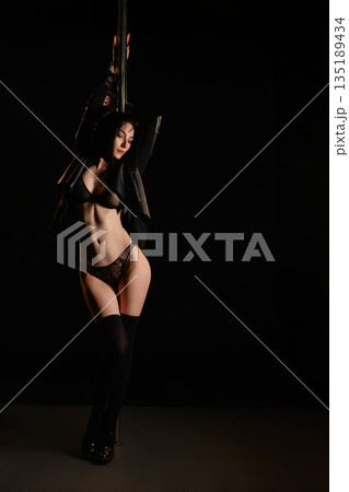 Sensual woman in black lingerie and jacket posing with dance pole in dark studio. 135189434