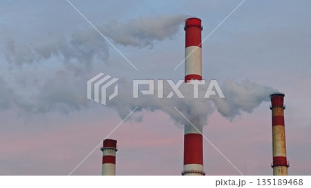 Industrial chimneys piercing cloudy sky. Tall red and white striped factory smokestacks emerge from low clouds against blue and pink sky. Industrial chimneys piercing cloudy sky. Tall red and white striped factory smokestacks emerge from low clouds against blue and pink sky. 135189468