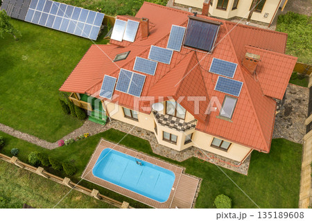 Aerial view of a new autonomous house with solar panels and water heating radiators on the roof and green yard with blue swimming pool. 135189608