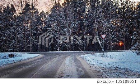 Snowy forest road with yield sign at sunset. Winter scene of snow-covered trees along empty road with triangular yield traffic sign under pink sunset sky. Snowy forest road with yield sign at sunset. Winter scene of snow-covered trees along empty road with triangular yield traffic sign under pink sunset sky. 135189651