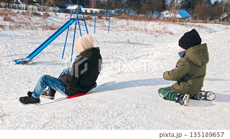 Children sled on snowy playground. Two children sit on red sled near blue slide on snowy playground with trees. Children sled on snowy playground. Two children sit on red sled near blue slide on snowy playground with trees. 135189657