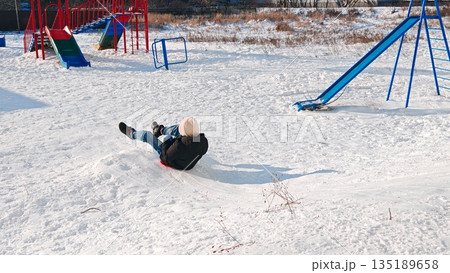 Child slides down snow mound. Child slides on red sled down snow mound at colorful playground. Child slides down snow mound. Child slides on red sled down snow mound at colorful playground. 135189658