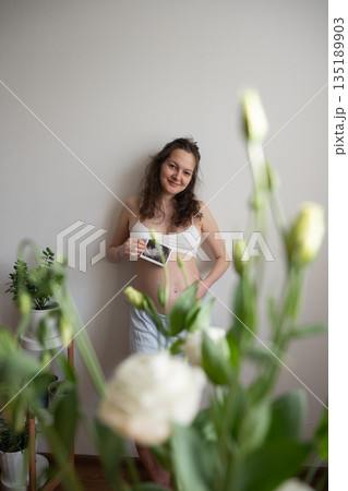 Pregnant woman showing ultrasound scan while standing next to houseplants Pregnant woman showing ultrasound scan while standing next to houseplants 135189903