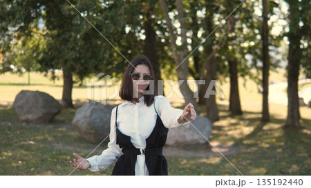 A business woman stands confidently in a park wearing sunglasses and a stylish outfit. She is gesturing as if explaining something important while surrounded by trees and rocks. A business woman stands confidently in a park wearing sunglasses and a stylish outfit. She is gesturing as if explaining something important while surrounded by trees and rocks. 135192440