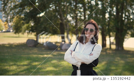 A professional woman stands smiling with her arms crossed in a sunny park. She wears stylish sunglasses and a chic outfit, enjoying a pleasant day surrounded by nature. A professional woman stands smiling with her arms crossed in a sunny park. She wears stylish sunglasses and a chic outfit, enjoying a pleasant day surrounded by nature. 135192442