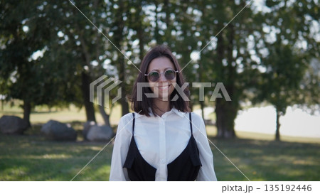 A confident woman stands outdoors in a park, wearing sunglasses and a stylish outfit. She smiles while surrounded by trees and natural beauty, enjoying a sunny day. 135192446