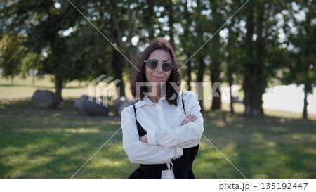 A woman stands confidently in a park, smiling with arms crossed. She wears stylish sunglasses and a black-and-white outfit, enjoying a pleasant day outdoors in a natural setting. 135192447
