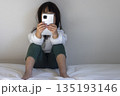 うす暗い部屋で座ってスマートフォンを見る女の子のイメージ（5歳、日本人、顔無し） 135193146