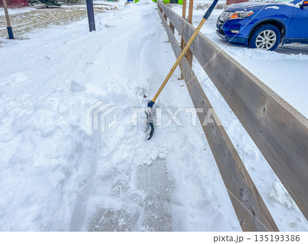 Snow shovel stuck in deep snow beside cleared icy sidewalk near wooden fence, winter maintenance concept, cold weather, snowfall aftermath, pedestrian safety and snow removal. 135193386