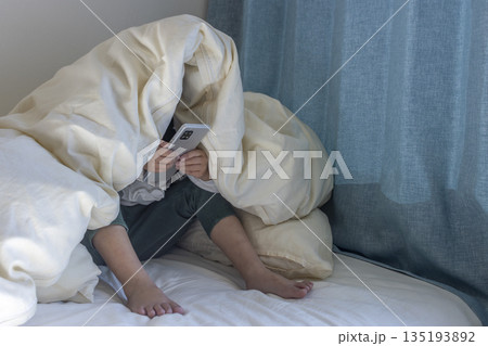 うす暗い部屋で布団をかぶってスマートフォンを見る女の子のイメージ（5歳、日本人、顔無し） 135193892