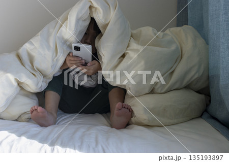 うす暗い部屋で布団をかぶってスマートフォンを見る女の子のイメージ（5歳、日本人、顔無し） 135193897