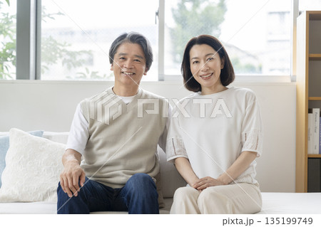 ソファに並んで座るカメラ目線で笑顔の50代の夫婦 イメージ ソファに並んで座るカメラ目線で笑顔の50代の夫婦 イメージ 135199749