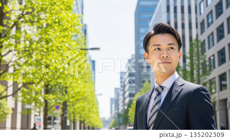 オフィス街で爽やかに微笑む日本人ビジネスマン｜新緑の背景とスーツ姿の男性・採用やキャリアのイメージ 135202309