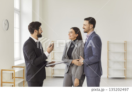 Smiling realtor presenting modern property to young couple and discussing purchase details together. Smiling realtor presenting modern property to young couple and discussing purchase details together. 135202497