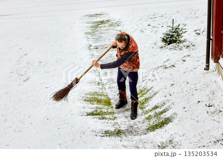 Adult person clears snow using long wooden broom while standing outdoors on winter ground captured from elevated angle during ongoing work. Adult person clears snow using long wooden broom while standing outdoors on winter ground captured from elevated angle during ongoing work. 135203534