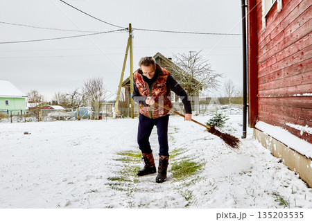Homeowner clears snow from garden path using broom wearing warm vest and winter boots. 135203535