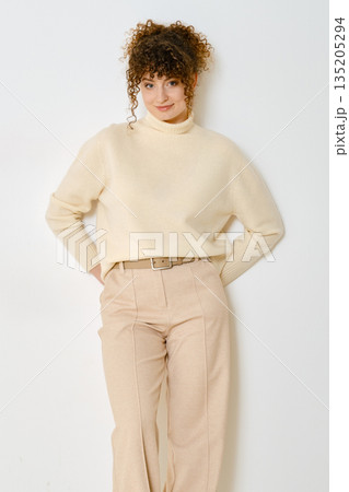 Young woman with curly hair poses in cream sweater and beige pants with a neutral background 135205294