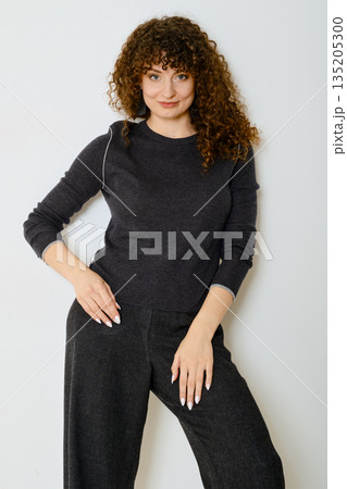 Tricky woman in dark sweater and matching trousers stands gracefully against white wall 135205300