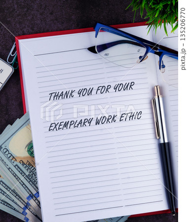 An open notebook displays a handwritten thank you message on lined paper. A pen, glasses, and stack of cash sit nearby on a dark surface 135206770