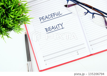 An open notebook has the handwritten phrase PEACEFUL BEAUTY on its lined pages. A pen, eyeglasses, and a small green plant are also arranged nearby 135206794