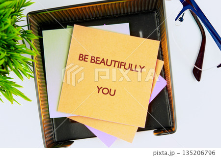 Close-up captures a note that reads BE BEAUTIFULLY YOU in a dark metal box. Colorful papers are underneath and a plant is sitting next to the box. A pair of glasses rests nearby 135206796
