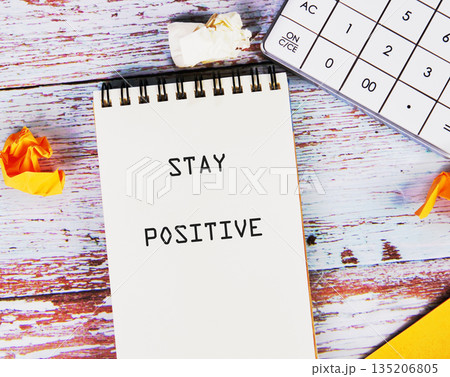A spiral notebook with the inspiring words STAY POSITIVE is on a weathered wooden table, next to a calculator and crumpled paper, creating a serene workspace A spiral notebook with the inspiring words STAY POSITIVE is on a weathered wooden table, next to a calculator and crumpled paper, creating a serene workspace 135206805