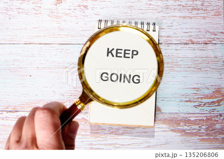 A hand holds a magnifying glass over a notepad. The notepad reads KEEP GOING. The background is a faded wooden surface. It encourages continuous effort and perseverance A hand holds a magnifying glass over a notepad. The notepad reads KEEP GOING. The background is a faded wooden surface. It encourages continuous effort and perseverance 135206806